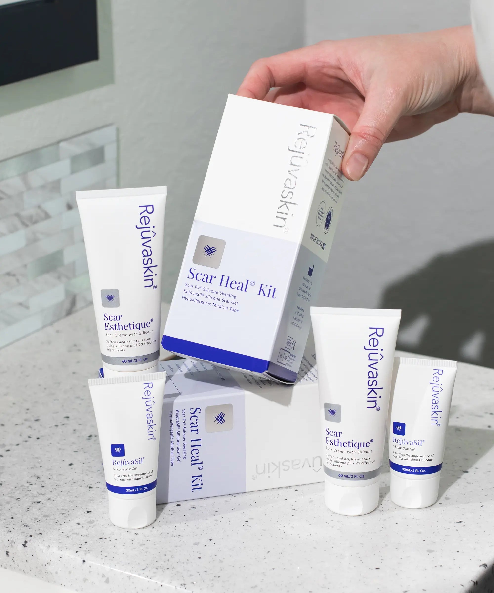 A hand holds a white and blue box of Rejuvenskind Scar Heal Kit, surrounded by four white tubes and two boxes of the same kit on a speckled white countertop.