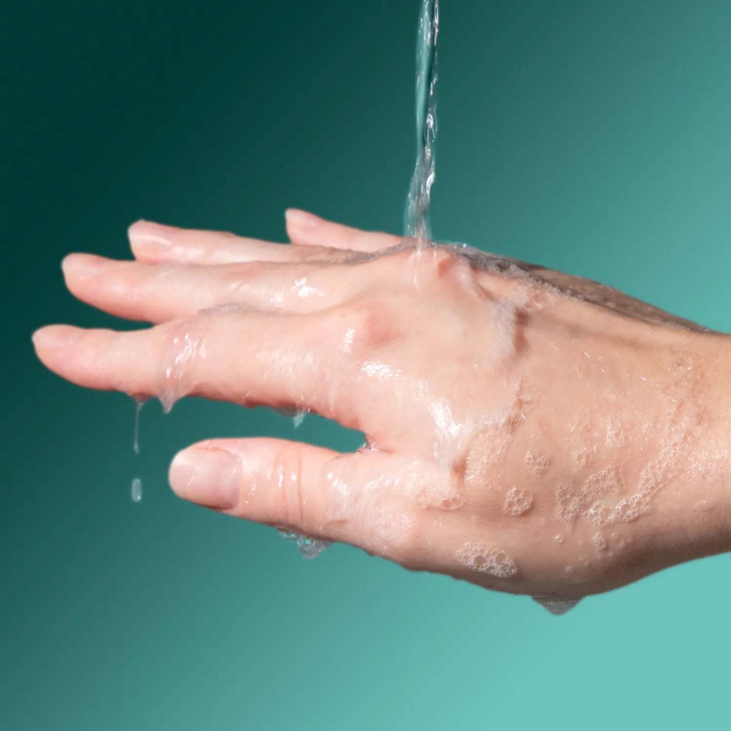 A close-up of a hand under running water with soap lather, shown against a teal gradient background.