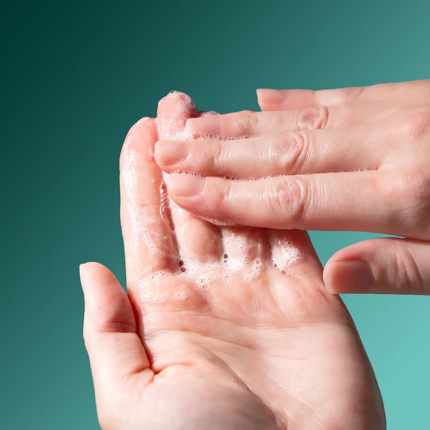 Close-up of two hands washing together with white soap foam, set against a solid teal background.