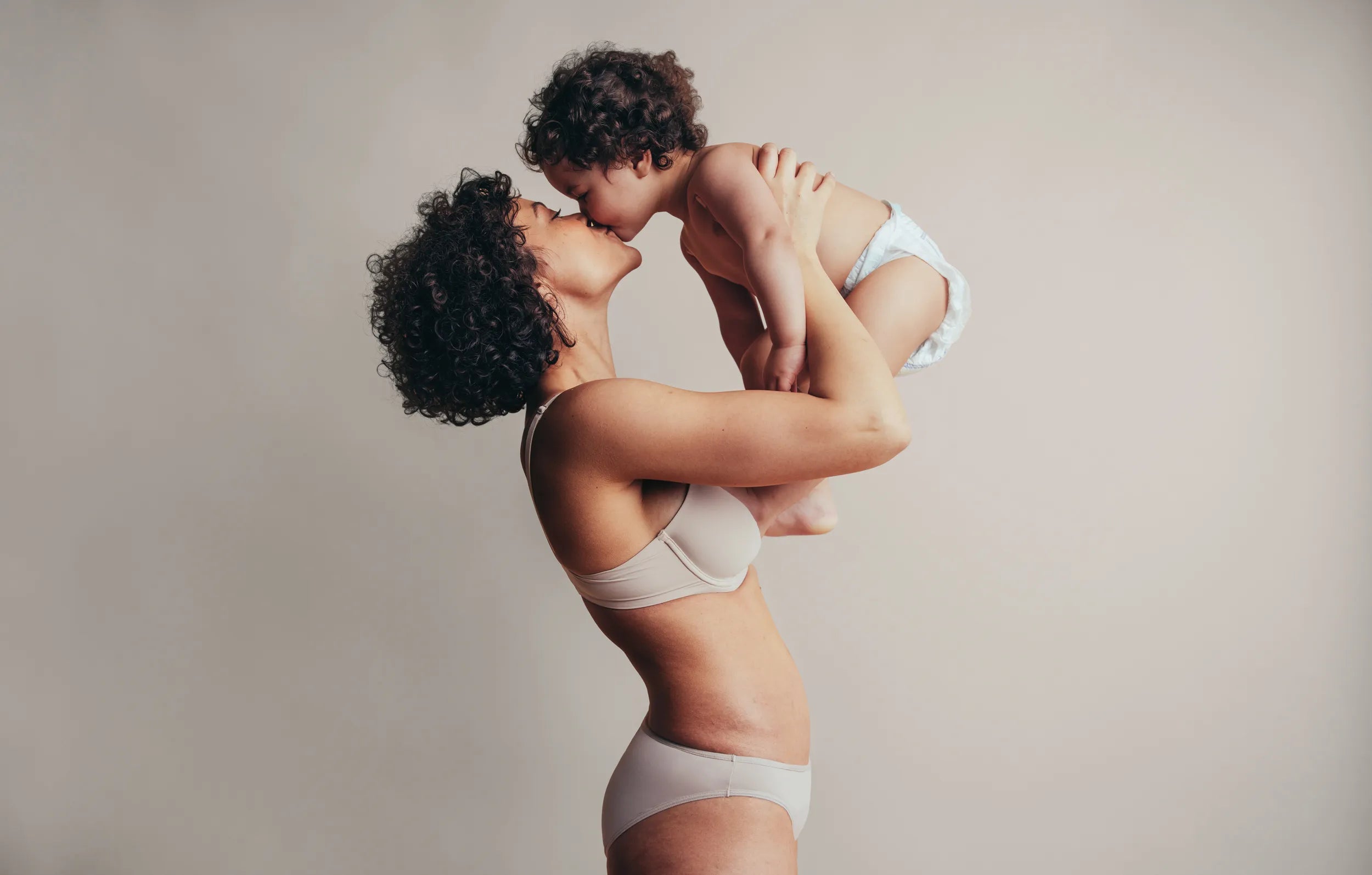 A woman in a light beige bra and underwear holds a young child in a white diaper against a plain beige studio background.