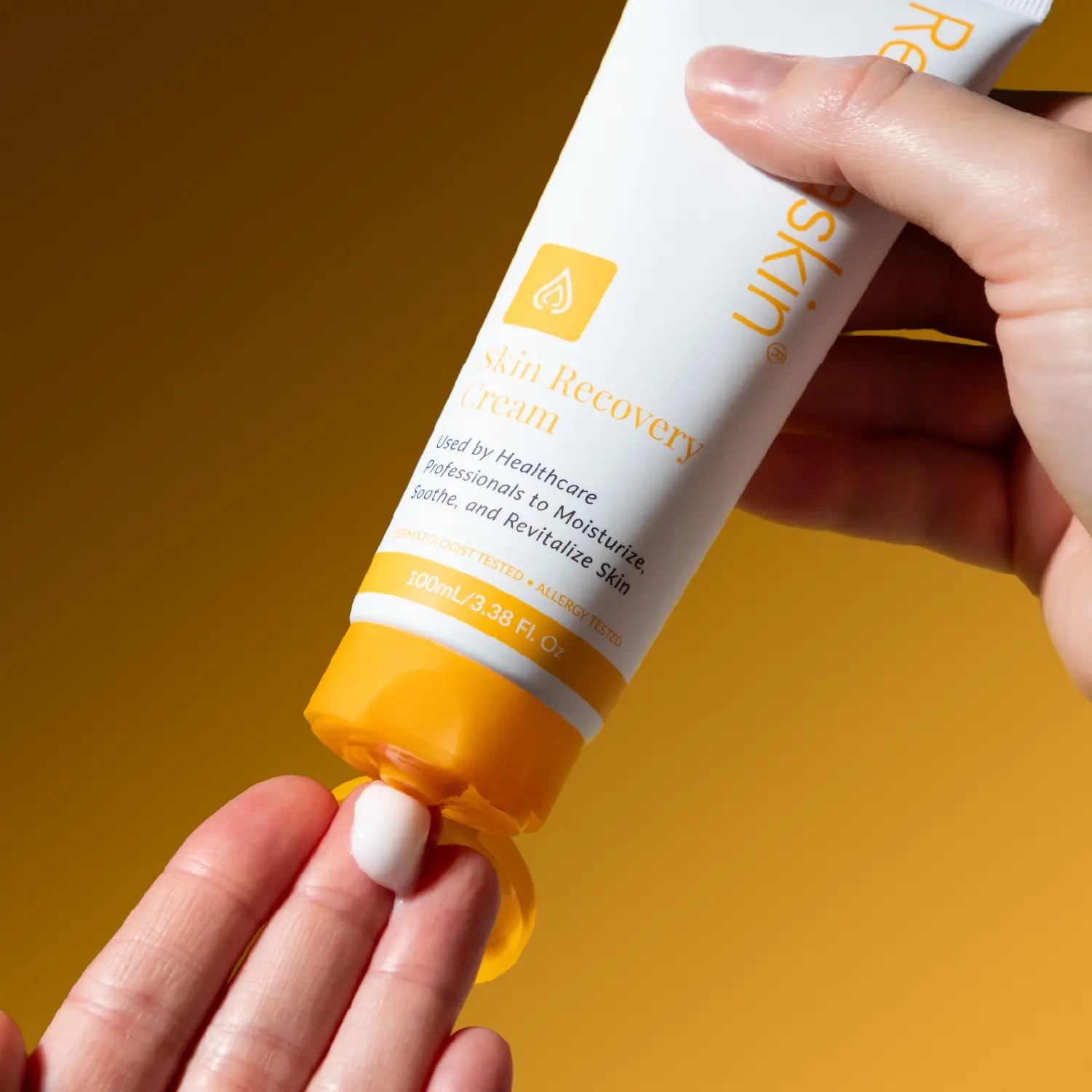 A hand holds a white and yellow tube of Re:Skinned Skin Recovery Cream, squeezing a small white dollop onto another hand against a warm yellow background.