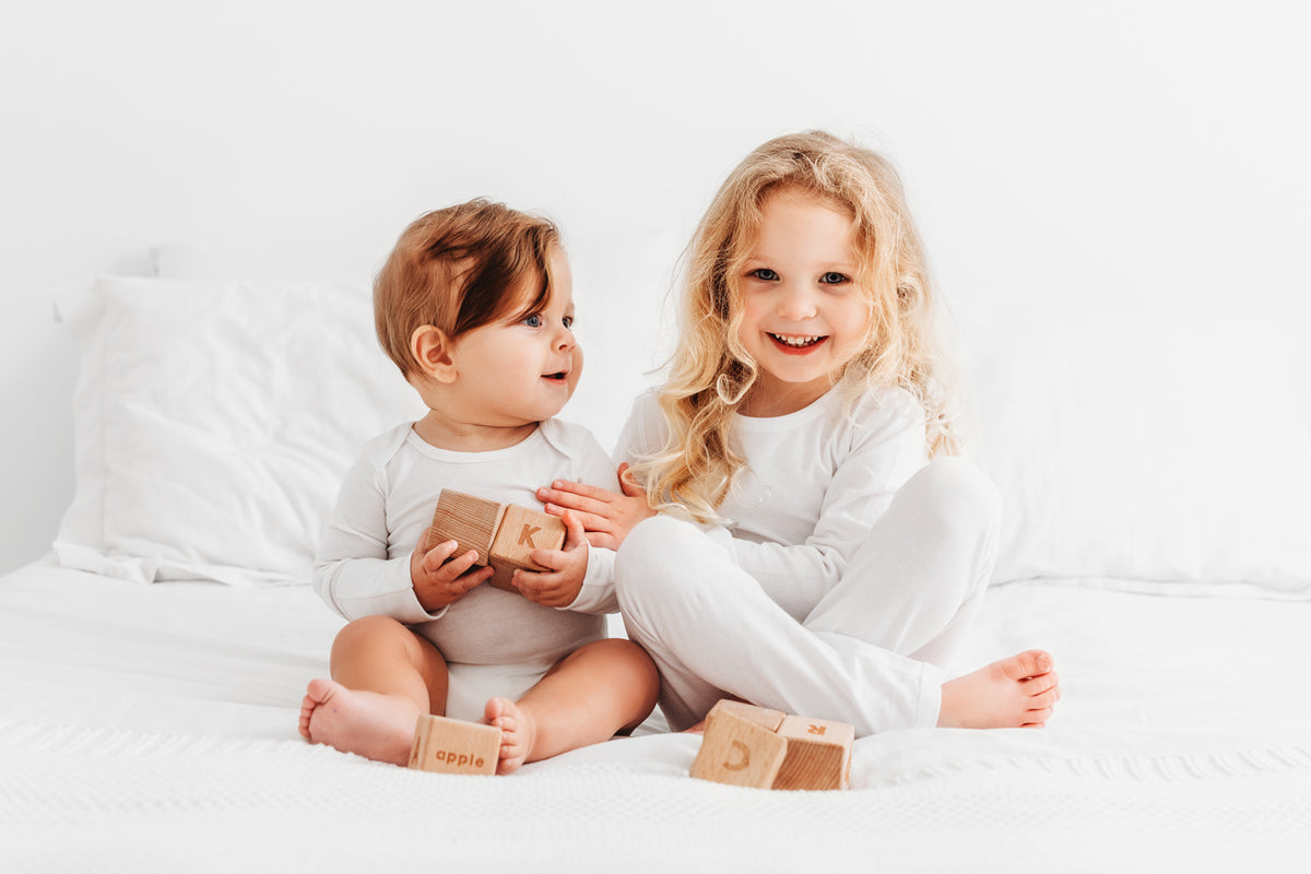 Two young children sitting on a white bed, wearing matching white long-sleeve pajamas and playing with small wooden blocks.